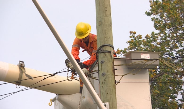 A man in a yellow hardhat and orange jumpsuit stands in the bucket of a crane and works on wires.