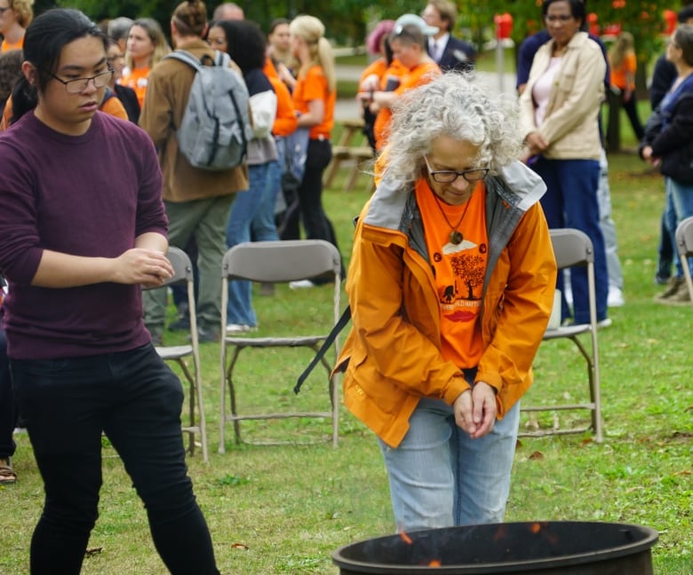 A person dressed in orange throws a handful of tobacco into a firepit as a line forms behind them. 