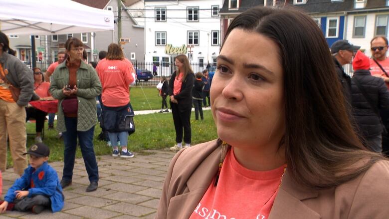 A woman wearing a beige blazer and orange shirt stands in a public park.