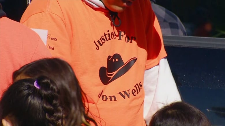 A man wears an orange t shirt with 'Justice for Jon Wells' written on it. 