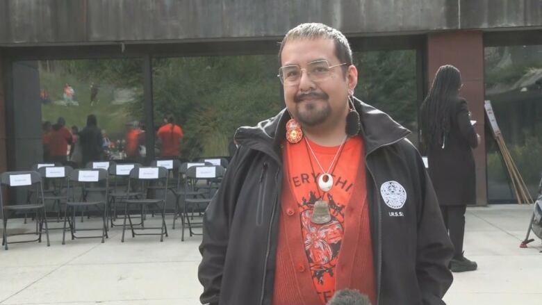 A man wearing an orange shrit and a black jacket smiles in front of a building.