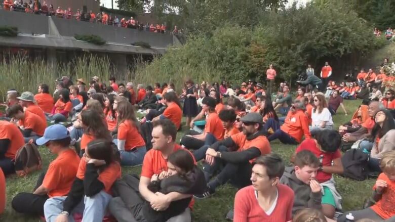 A sea of people wearing orange listen to a speaker while seated on a lawn.