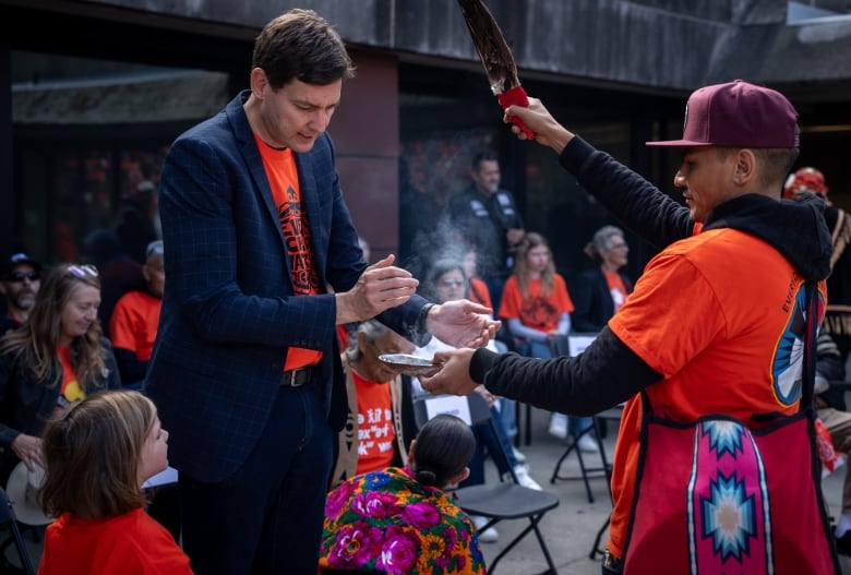 A man in a blue suit and orange shirt smudges smoke from a tray held by a man with an eagle feather.