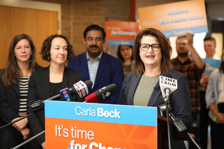 A woman in a green shirt and a dark blue blazer stands behind a podium. A sign on the podium identifies the woman as Carla Beck. Behind her are people waving signs while two women and a man in formal clothing stand to the left of the podium.