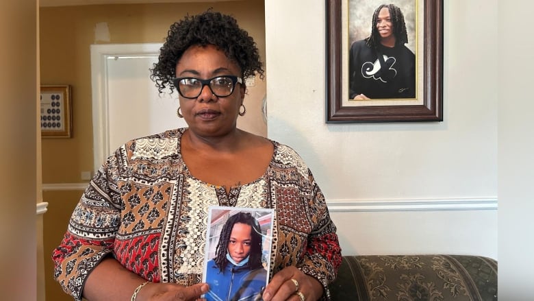 A woman holding up her son's photo and standing in front of a another photo of him on her wall. 