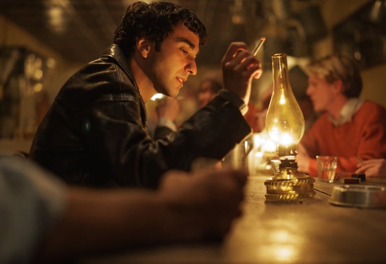 A man in a leather jacket sits at a dimly lit bar with a cigarette in hand.