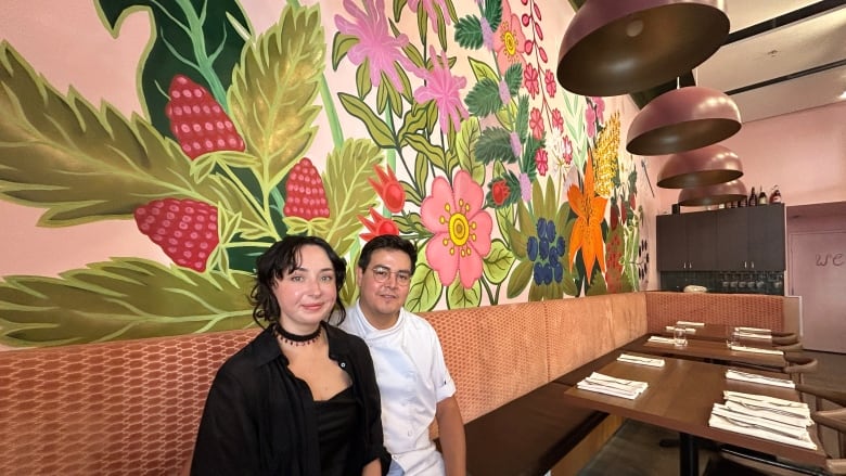 A woman and a man sit next to each other on bench seating. He wears chef whites, and behind them is a mural with flowers and raspberries.