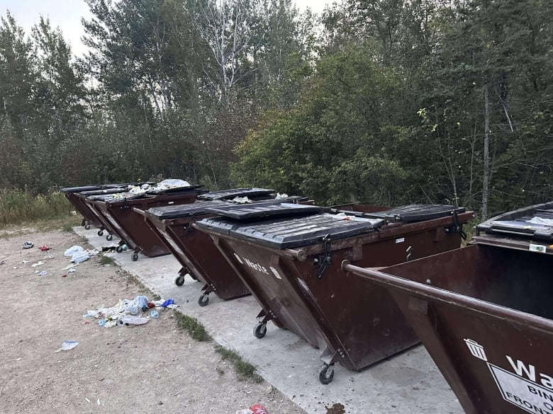 Five brown trash bins in a row have garbage strewn around it. 