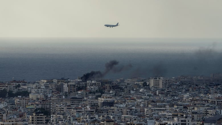 A commercial passenger plane flies over a city as smoke rises from a building below.