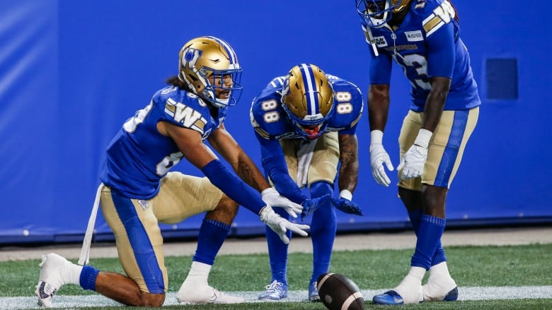 Three football players on the same team crouch on the ground near the football after their team got a touchdown. 