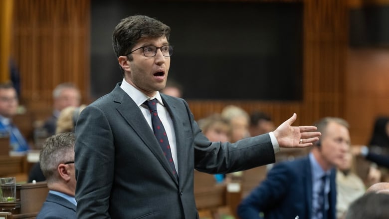 A man in a grey suit gestures with his left hand as he stands and speaks in the House of Commons.