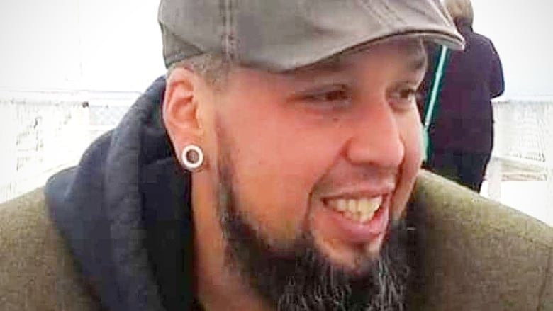 An Indigenous man with a long black beard, thin moustache, silver earring and wearing a heavy jacket and denim cap smiles.