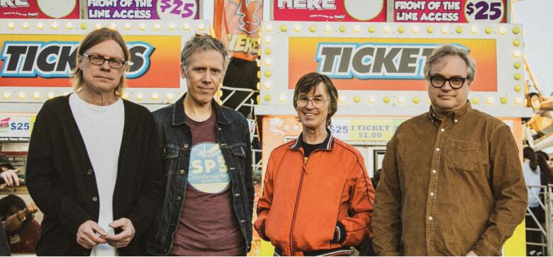 Four men standing in a row against a backdrop of signs that say Tickets and Front of the Line Access $25.