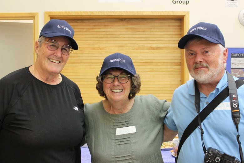 Three people smile at the camera wearing navy ball caps that say 