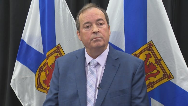 Man in blue suit and lavendar shirt and tie stannds in front of two Nova Scotia flags.