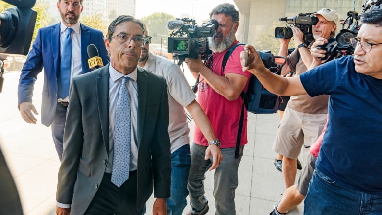 A man in a suit walks past a crowd of reporters.