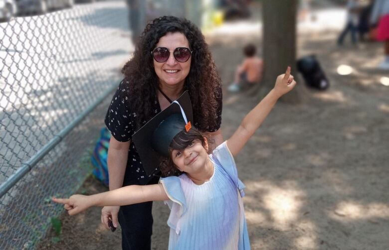 A woman with brown curly hair and sunglasses stands in front of a small girl with a graduation cap on. 