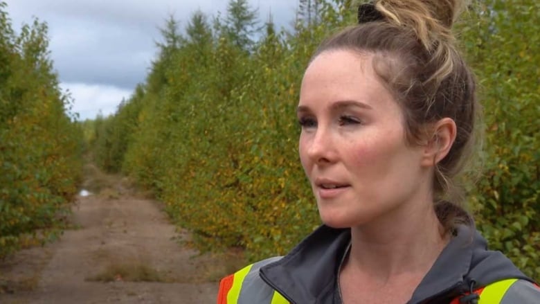 A portrait of a woman with her hair tied in a bun at the top of her head, wearing a reflective vest on a dirt road in the woods.