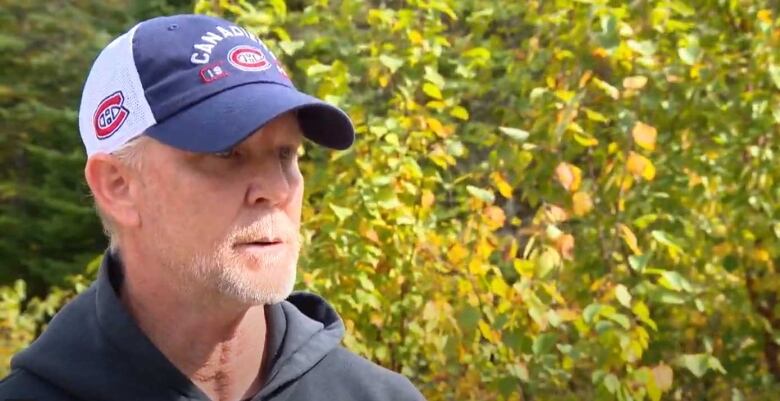 A portrait of a man with a greying goatee, wearing a hooded sweatshirt and baseball hat outside.