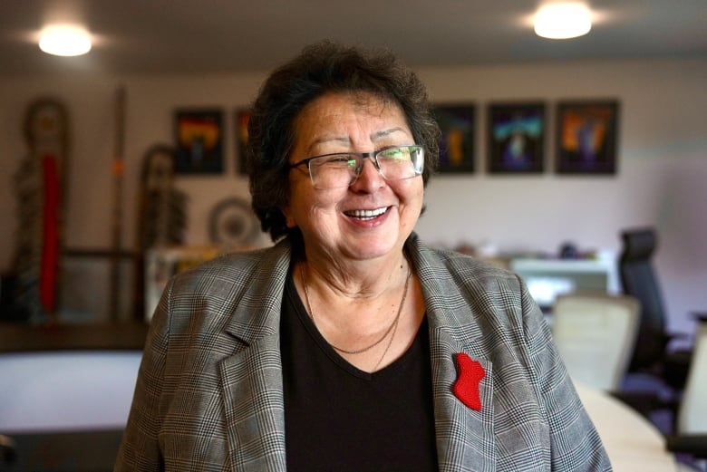 A woman wearing a black shirt and grey blazer with a red dress pin poses for the camera.