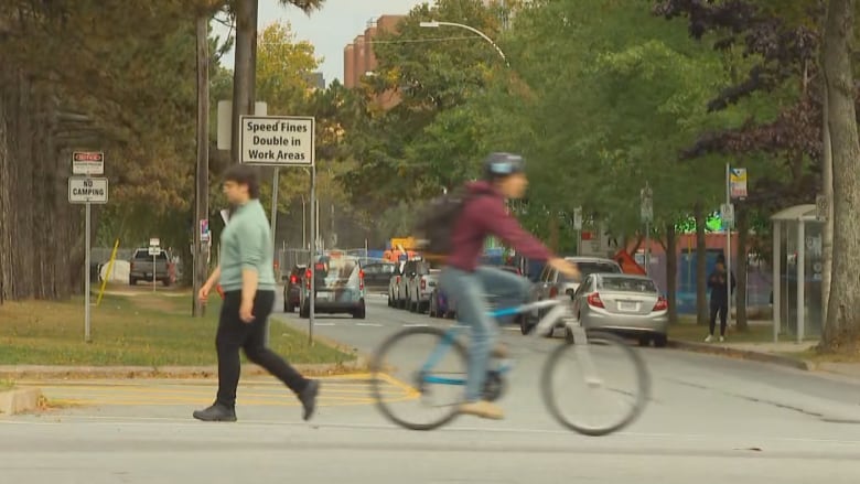 A cyclist passes a pedestrian at a crosswalk. Trees can be seen in the background.