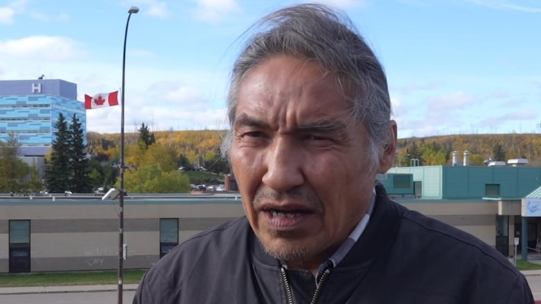 Indigenous man, in his late fifties, pictured from the chest up, hair blowing over his head. A road and buildings in the background. 