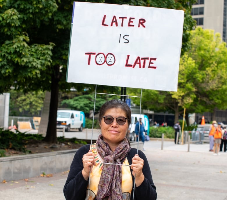 Woman with sunglasses holds sign that says 'Later is too late.'