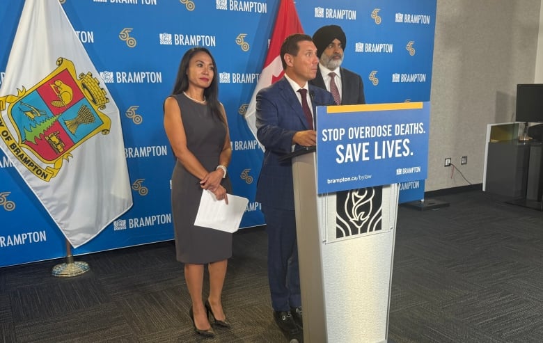 Two men in suits and a woman in business attire stand at a podium inside with a Brampton banner and flag in the background