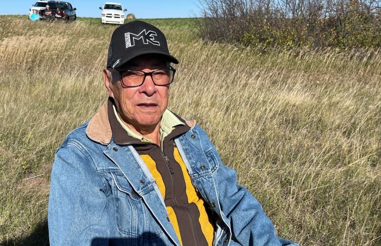 A man wearing a hat and denim jacket sits in a field of grass. 