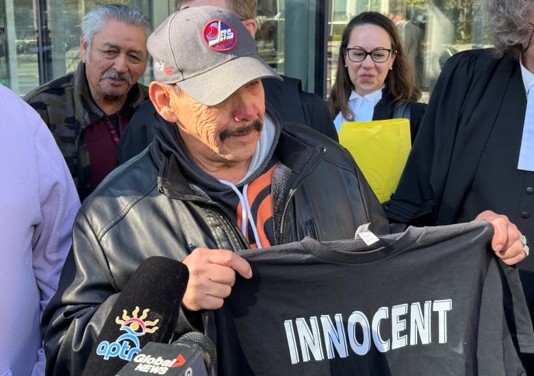 A man wears a baseball cap and holds a T-shirt that has the word 