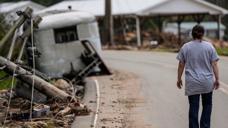 A person walks down a road, their back to the camera. Rubble is seen at the side of the road, including what looks like a vehicle. There is a building in the distance. 