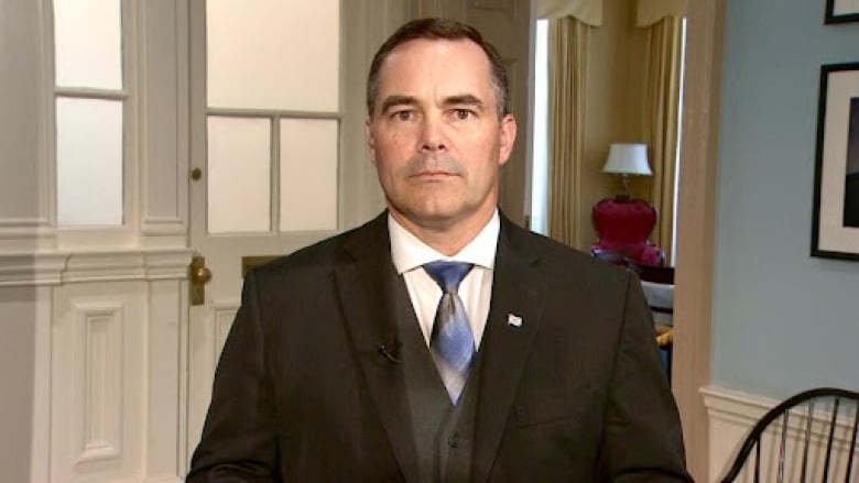 A man in a dark suit with a white shirt and blue and white tie stands with a neutral expression.