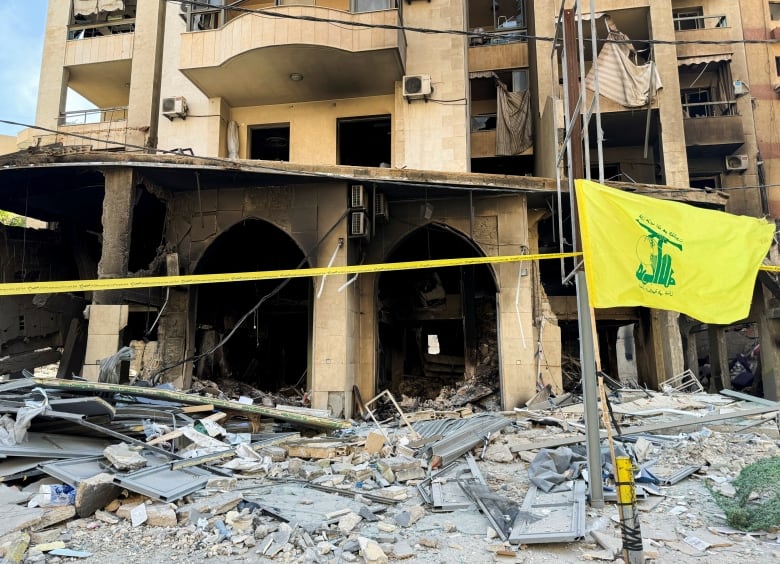 Concrete and metal debris are strewn across the ground in front of a heavily damaged multi-storey building with windows blown out. Nearby, a yellow flag with green detailing is shown.