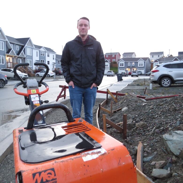 A man standing next to an orange painted piece of equipment, Behind him is rubble and a cul-de-sac lined with homes.