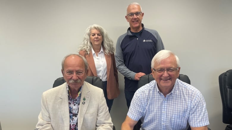 Four people are looking are smiling at a camera. Two people in the front are sitting at a desk and holding pens.