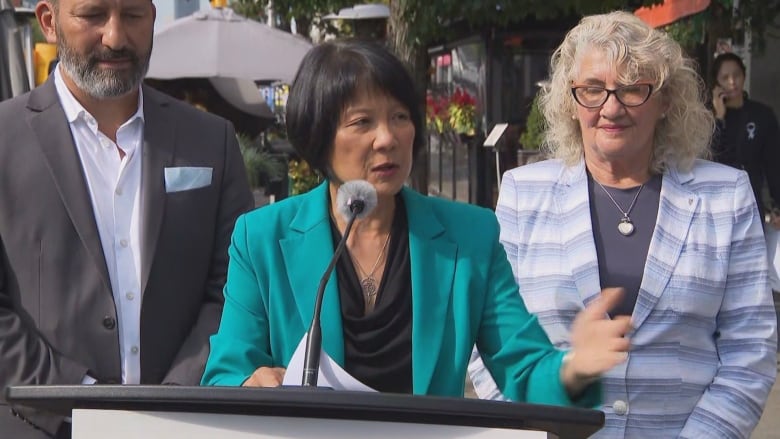 Toronto's mayor speaks into a microphone at a podium outside on a sunny fall day on a city street. A man and woman stand behind her on either side. She's visible from the waist up.