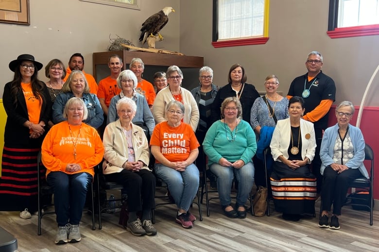 A group of people stand and sit next to each other. Many are wearing bright orange shirts.