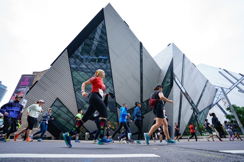 On an overcast fall day, dozens of runners with racing bibs run on a city street passing the glass, Royal Ontario Museum