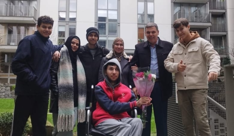 A man and a woman stand smiling, surrounded by five teenagers, including one young man sitting in a wheelchair and holding a bouquet of flowers.