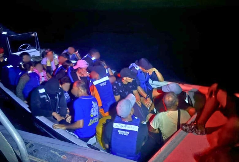 People whose faces have been blurred are seen wearing blue personal floatation devices crowd in a small boat on the ocean at night. 