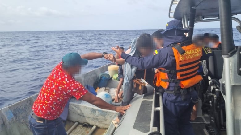 A man in a red shirt and ball cap stands on a small boat and helps a woman into a larger vessel into the arms of a Colombian Navy official.