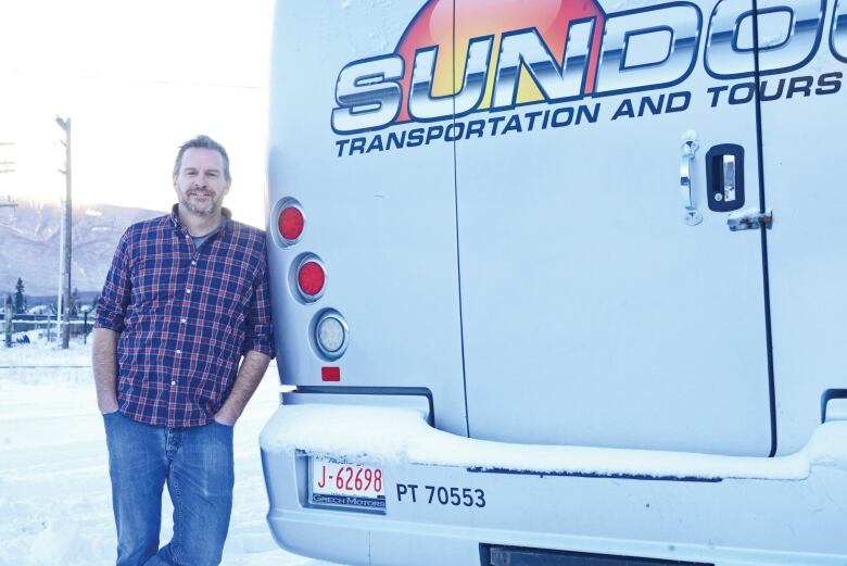 A man wearing a plaid shirt leans against a white van outdoors, smiling at the camera.