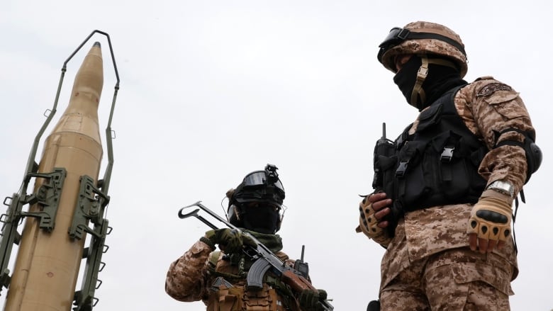 Two soliders, their faces in shadow, stand next to a towering missile.