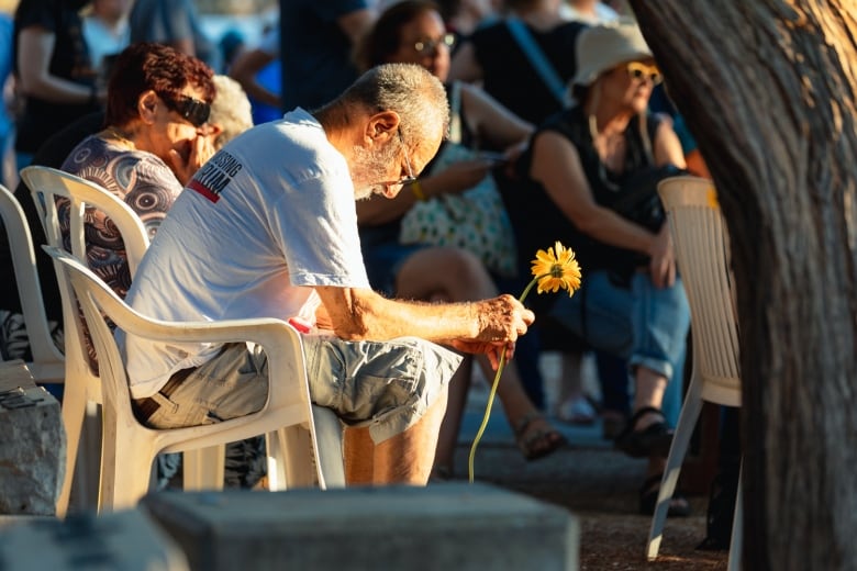 A seated person bows their head while holding a sunflower during a funeral.