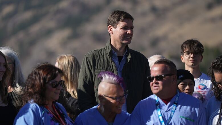 A tall white man smiles as people in scrubs talk underneath him.