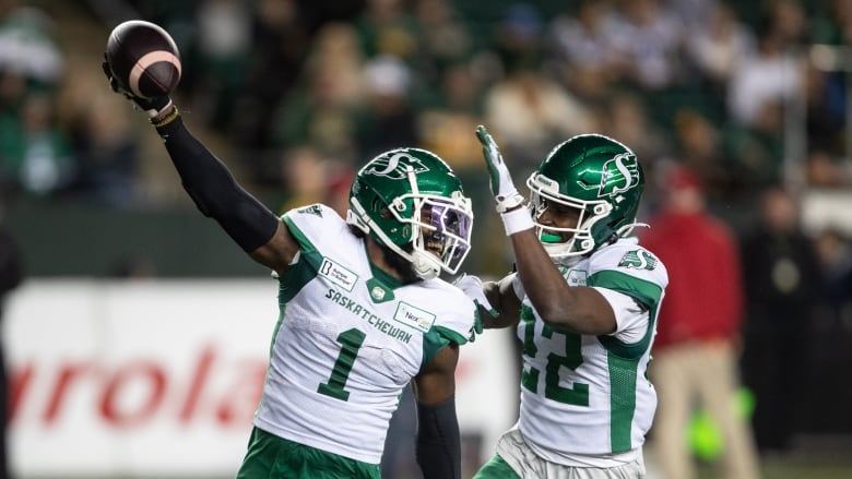 Two football players in white-and-green uniforms are cheering on the field. One of them is running while holding a football up in the air with their right hand.