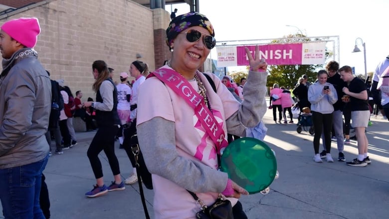 A woman wearing a bandana, sunglasses and a sash holds a peace sign to the camera.