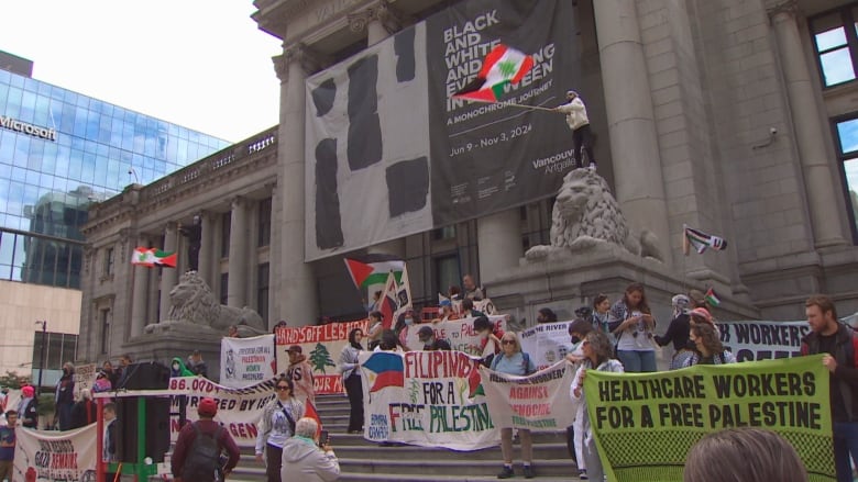 A demonstration outside a building with dozens of people holding colourful signs and waving flags.