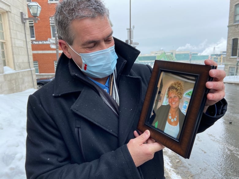 A man holds a framed picture of a woman and wears a mask with a small red 