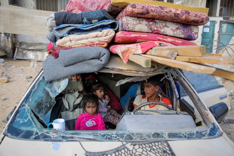 A family sits in a car with a missing windshield and stacks of blankets and mattresses strapped to the roof. 